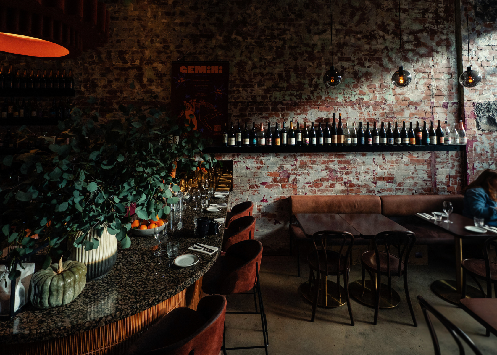 Dining area with brick wall, bar counter, and patrons in a cozy wine bar setting.